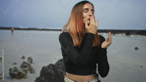 Foto Woman blowing kiss with hands toward camera on rocky beach shoreline at low tide, closed eyes and puckered lips; affection greeting farewell