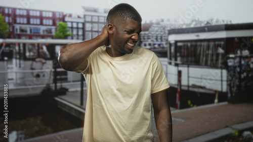 Εκτύπωση καμβά Man holding neck for backache and grimacing on street by a canal boat and dock, with nearby buildings and pavement visible; discomfort