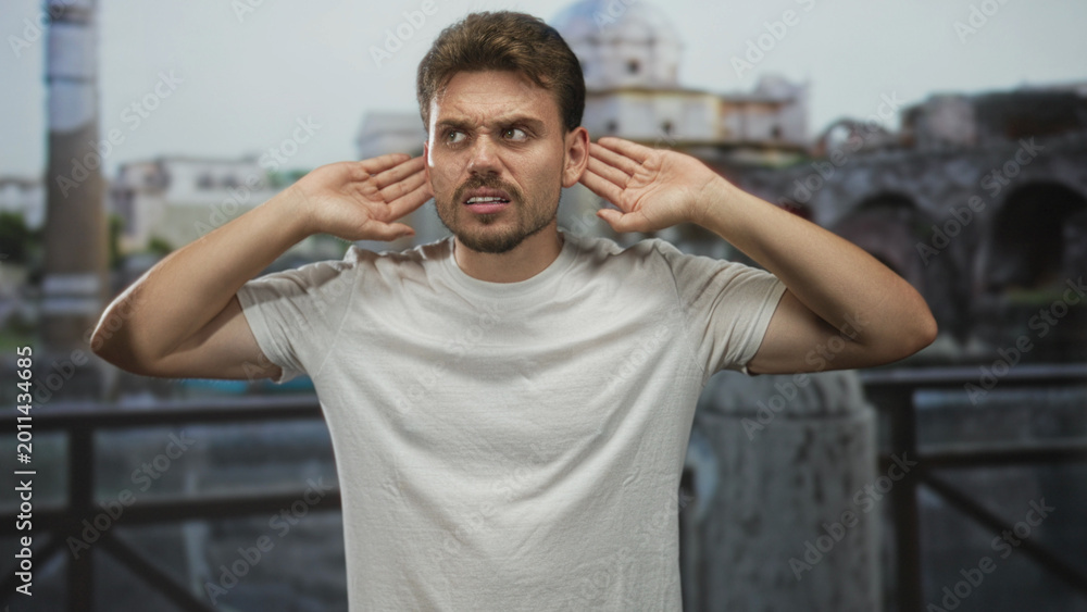 Fototapeta premium Man with hands cupping ears at building ruins on an outdoor balcony, looking upward and tensing shoulders; frustration focus.