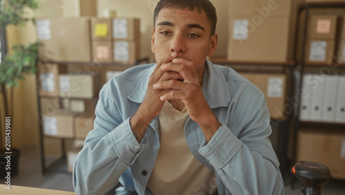 Bild auf Leinwand Man holding red smartphone with hands on desk, glancing down and clasping fingers, boxes stacked on shelves behind him in building; concern focus