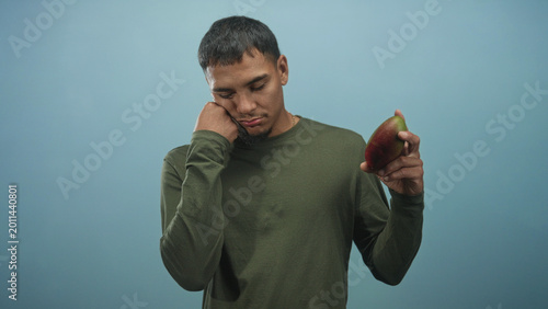 Schilderij op canvas Man in olive long sleeve shirt resting cheek on closed fist, holding a ripe mango in front of a soft blue studio backdrop; melancholy reflection