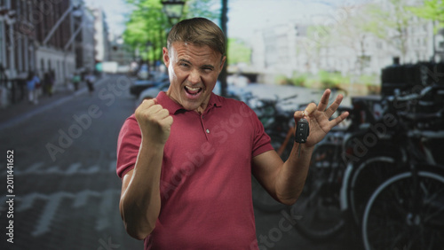 Schilderij op canvas Man in red polo holds car keys aloft with a clenched fist, grinning while pumping his arm on a street lined with parked bicycles and canal buildings; triumph success