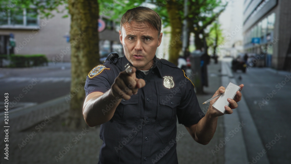 Fototapeta premium Police officer man pointing finger and holding notepad with pen on urban street by trees and building, badge and radio visible; seriousness duty.
