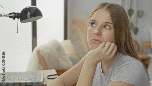 Tableau sur toile Young woman resting hand on cheek at study desk with lamp and hardcover book, gazing upward with pensive expression in a cozy home nook; contemplation