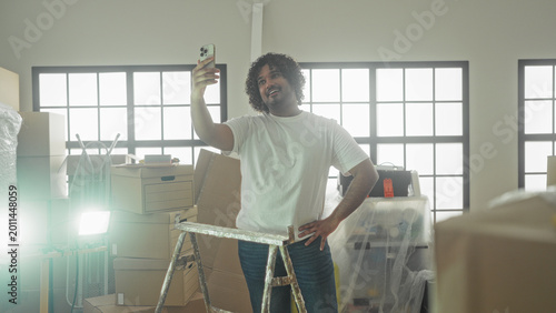 Foto Man holds smartphone for selfie with hand on hip amid packed moving boxes and stepladder in building; new home pride