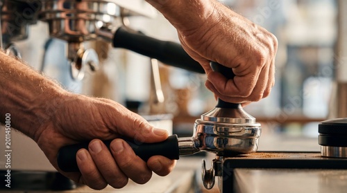 Barista tamping fresh ground coffee in a portafilter using a professional metal tamper in a cafe.