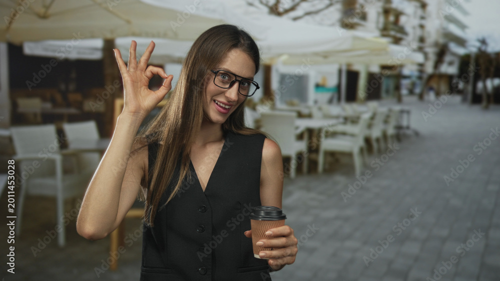 Fototapeta premium Woman making ok sign with hand and holding takeaway coffee cup on street cafe terrace at a restaurant with glasses and smile; joy confidence casual.