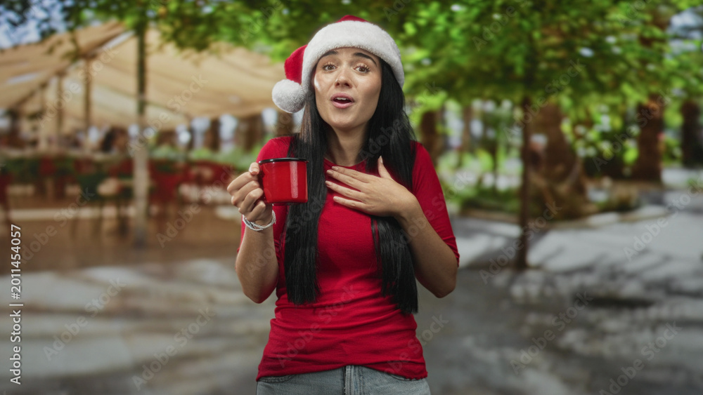 Fototapeta premium Young beautiful hispanic woman in santa hat holds red mug, hand to chest and eyes closed in a street cafe with canopy and trees; holiday joy.