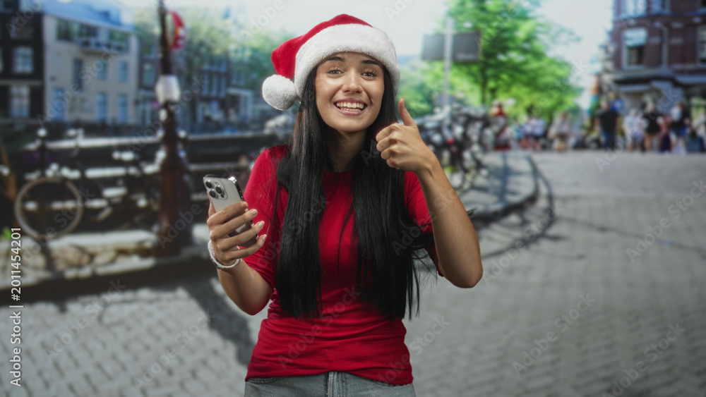 Obraz premium Woman wearing santa hat holding smartphone and giving thumbs up on cobblestone street by a canal with bicycles; festive joy.