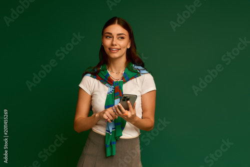 Fotografia Stylish young woman poses confidently in a green studio, holding her phone and s