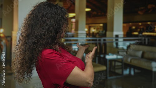 Cuadro en lienzo Woman with curly hair in a red shirt gesturing with bare arms in a hotel lobby; warm friendly welcome