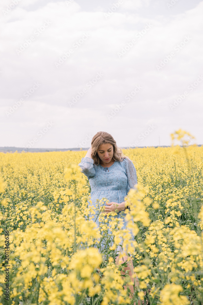 Fototapeta premium A beautiful pregnant woman with long wavy hair, wearing a light blue floral dress, gently holding her belly while standing in a blooming yellow rapeseed field. 