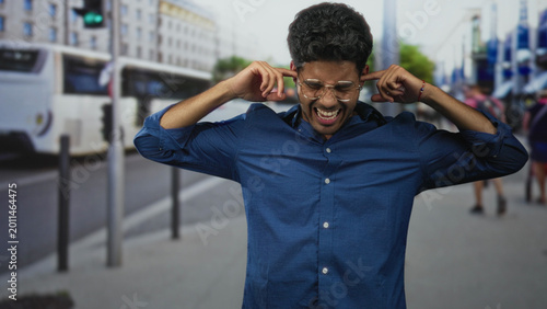 Foto Man in blue shirt and glasses pressing fingers into ears and grimacing on a busy city street beside a bus; stress overload