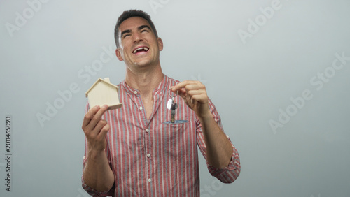Konstfotografi Young hispanic man smiling and holding a small wooden house and a set of keys in a pale blue studio; homeownership joy
