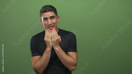 Billede på lærred Young hispanic man with hands covering mouth and tense expression in green studio; nervousness uncertainty