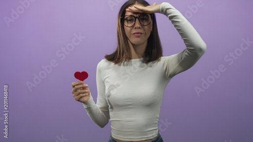 Foto Woman holding small red heart and shielding eyes with hand, wearing glasses in purple studio, frontal pose and visible hand gesture; hopeful longing