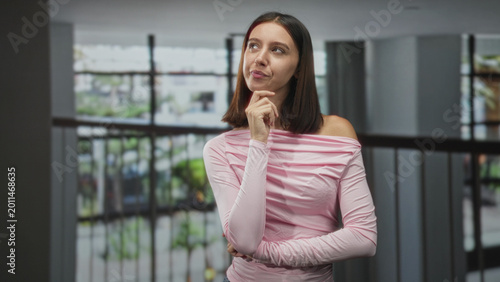 Bild auf Leinwand Woman with finger to chin and bare shoulder standing by railing in a modern building; thoughtful curiosity