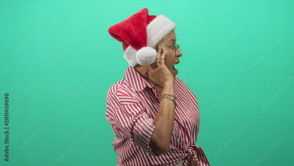 Naklejka premium Woman senior hispanic wearing santa hat cups ear with hand and open mouth, red striped shirt tied at waist against green studio backdrop; holiday surprise.