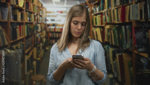 Fotografija Young blonde woman standing amidst bookshelves holding smartphone scrolling through messages in building; disappointment