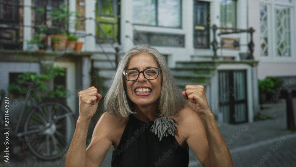 Obraz premium Grey haired woman with bare shoulders and arms, hands together above head on a cobbled street in front of a building and bicycle; joyful celebration freedom.