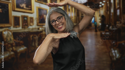 Quadro su tela Woman with bare arms and glasses raises hands framing smiling face in ornate gallery building corridor; joy celebration