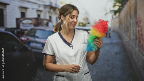 Canvastavla Woman cleaner holding rainbow feather duster with both hands, smiling while posed in a narrow street with parked cars; diligence care