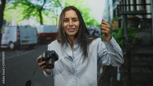 фотографія Woman holding camera with clenched fist and pointing gesture on street beside parked vans, face contorted in distress; frustration determination
