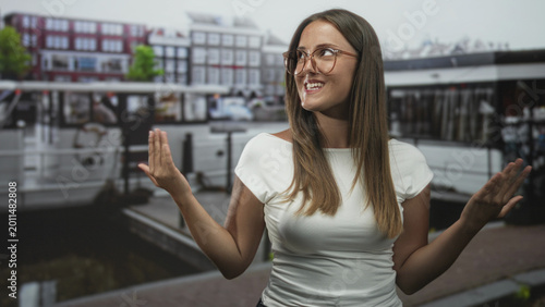 Bilde på lerret Woman shrugging with palms up and bare hands beside canal boats on a city street, smiling with closed eyes; playful ease