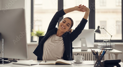 Happy Young Businesswoman in Black Blazer and White Blouse Stretching with Arms Overhead at a Modern Office Desk with Computers and White Coffee Cup