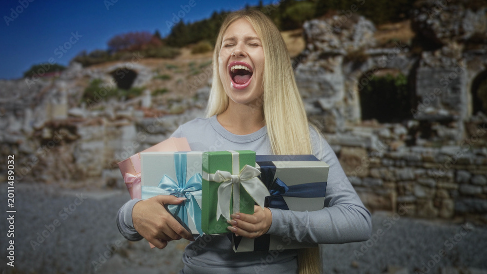 Obraz premium Woman clutching multiple wrapped gift boxes, mouth open as she shouts while standing by ancient stone building ruins; joy celebration.