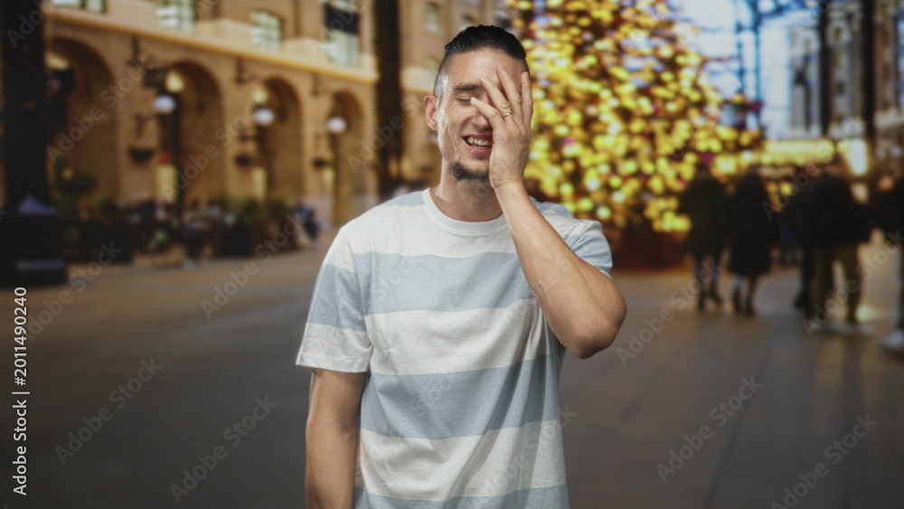 Fototapeta premium Man covering face with hand while smiling in a decorated building with blurred christmas tree and shoppers; embarrassed amusement.