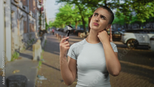Tableau sur toile Woman holding makeup brush and touching neck with a pained squint on a city street; self care discomfort