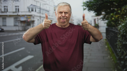 Konstfotografi Man giving double thumbs up on street in front of white townhouses, wearing maroon t shirt with bare forearms visible; encouragement optimism