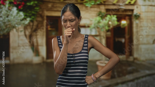 Bilde på lerret Thai woman touching forehead and coughing, wearing a striped tank top and bracelet, standing before a stone building entrance on a street amid potted plants; fatigue seeking help
