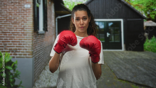 Tableau sur toile Woman wearing red boxing gloves holds fists up near chest in front of building entrance on a residential driveway, direct gaze and relaxed stance; confidence training