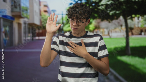 Foto Teenage boy wearing glasses and a striped shirt places hand on chest and raises other hand on street; sincerity pledge