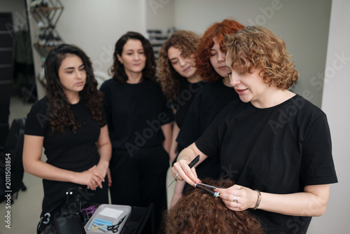 Professional hairdresser instructor teaching students biowave and permanent curling techniques using perm rods on a mannequin head during a beauty school masterclass.