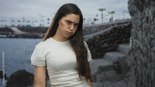 Foto Woman in white shirt touching cheek on seaside by coastal building under cloudy sky with stone wall; boredom