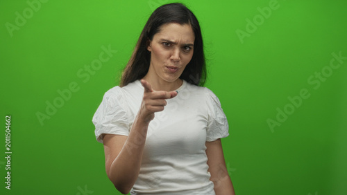 Fotografia Young hispanic woman pointing finger in green studio with serious expression and tense posture; anger