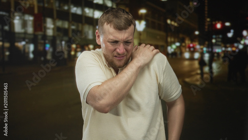 Fotografía Man clutching his shoulder and grimacing on a busy city street at night under streetlights and blurred cars; pain
