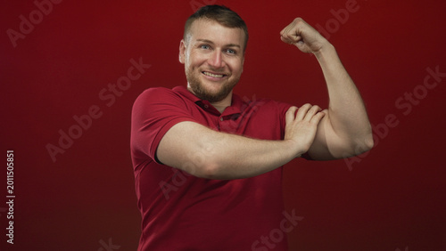 Valokuva Young heavyset man flexing biceps with clenched fists in a red studio set, smiling at the lens; confidence strength