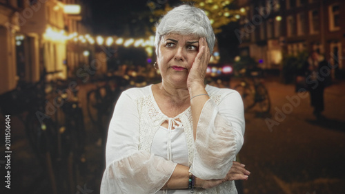 Foto Woman with grey hair and white blouse touching her face and crossing arms on a night street lined with bikes and string lights; tired reflection