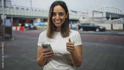 Bild auf Leinwand Woman smiling while texting on smartphone on street, holding phone with both hands and briefly giving thumbs up; everyday happiness
