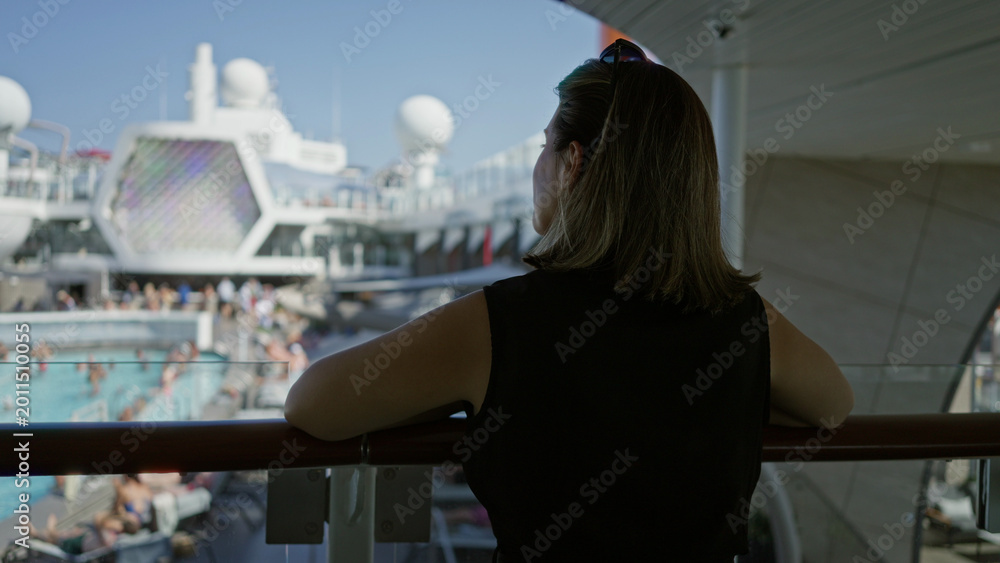 Obraz premium Woman leans on ship rail with arms resting on railing and looks over crowded pool on deck; calm contemplation.