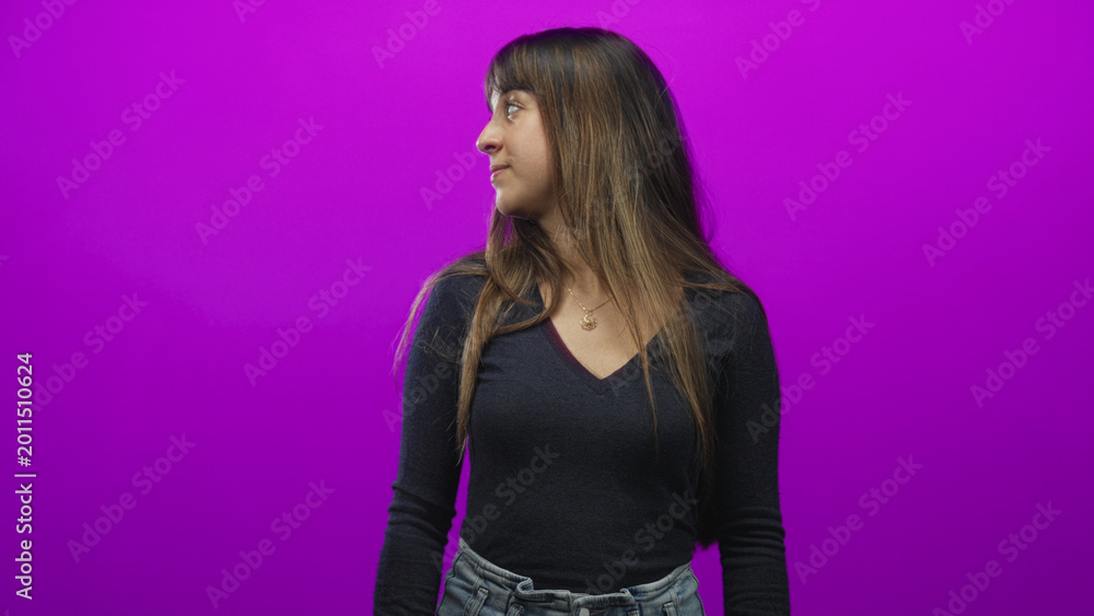Naklejka premium Young woman wearing pendant necklace and high waisted jeans turns head to side looking left against a vibrant purple studio backdrop; contemplation.