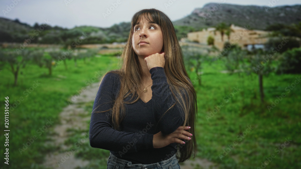 Obraz premium Woman with hand on chin and long hair covering face in front of building on a grassy path; quiet introspection.
