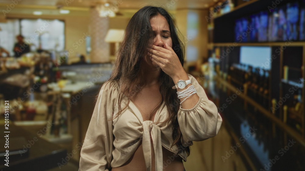 Fototapeta premium Hispanic woman clutching chest and covering mouth at a restaurant bar counter amid blurred patrons and bottles on shelves; distress nausea.