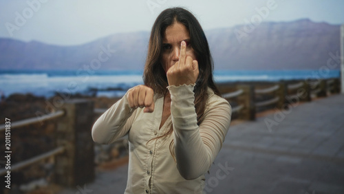 Bild auf Leinwand Young hispanic woman showing middle finger and clenched fists on a street promenade by the seaside; defiance