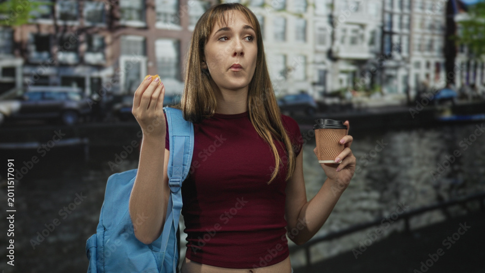 Fototapeta premium Student woman eating snack with hand to mouth and holding takeaway coffee cup on street, blue backpack visible; morning break contentment.