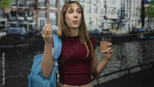 Foto Student woman eating snack with hand to mouth and holding takeaway coffee cup on street, blue backpack visible; morning break contentment